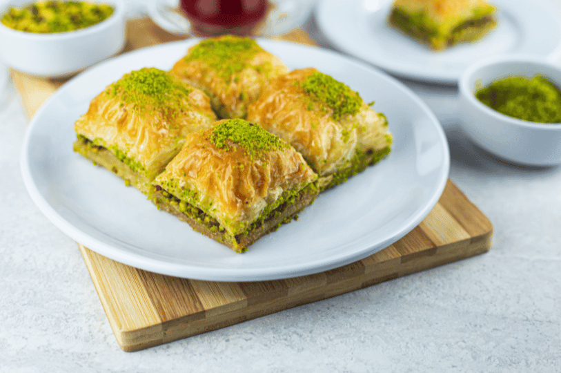 A bright, clean shot of four baklava squares on a white plate resting on a wooden board, with small bowls of extra pistachio garnish in the soft-focus background.