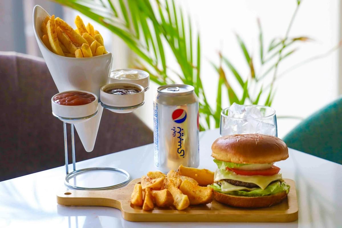 A beef burger topped with cheese, tomato, and lettuce is served on a wooden board alongside thick-cut wedges and a silver soda can. In the background, a modern metal stand holds a cone of french fries and three small bowls of dipping sauces.