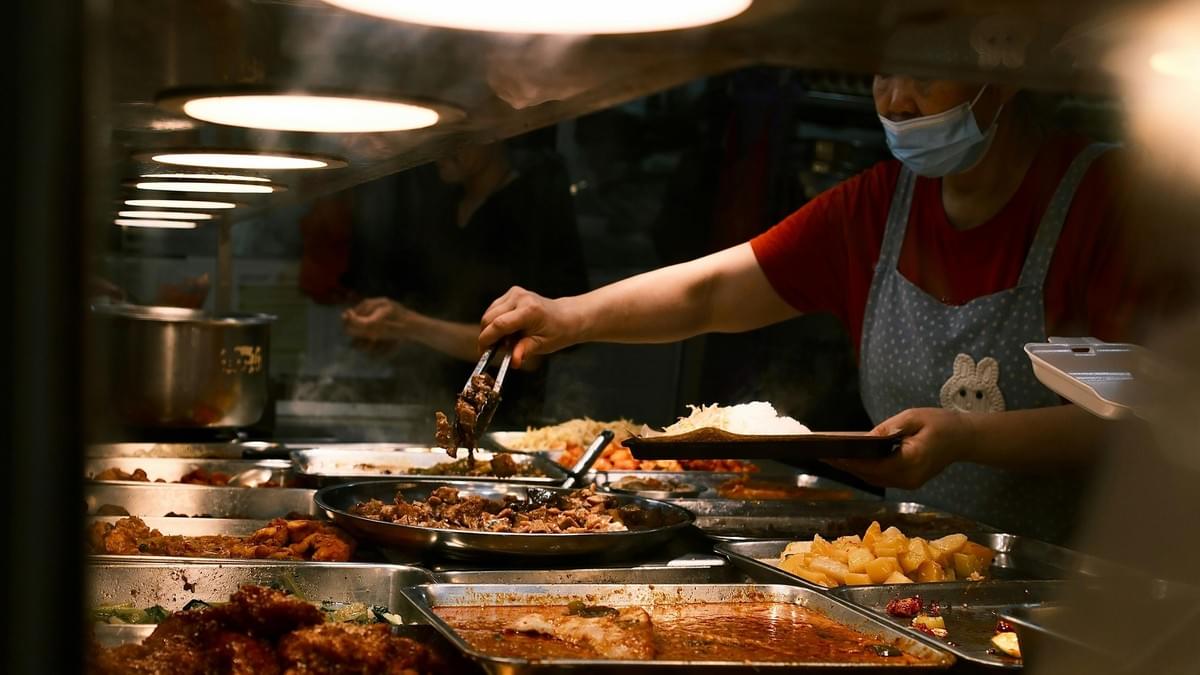 Captured from the perspective of a bed with rumpled white linens, this image frames a stunning city skyline through large, dark-framed windows. The sky transitions from a cool blue to a vibrant orange at the horizon, silhouetting the urban architecture during what appears to be a peaceful sunrise or sunset.