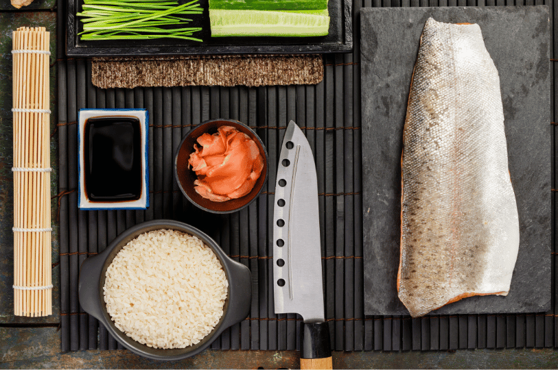 A curated arrangement of sushi-making essentials—knife, raw salmon, nori, rice, and vegetables—laid out on a black bamboo mat, evoking the seasonal precision and ingredient-forward ethos of Japanese dining in Singapore.