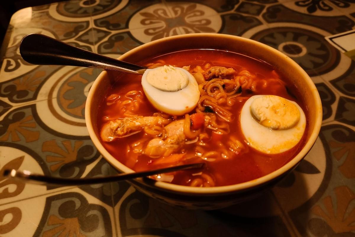 A bowl filled with spicy red ramen broth, noodles, and two hard-boiled egg halves rests on a patterned table. A dark spoon rests inside the dish under warm lighting.