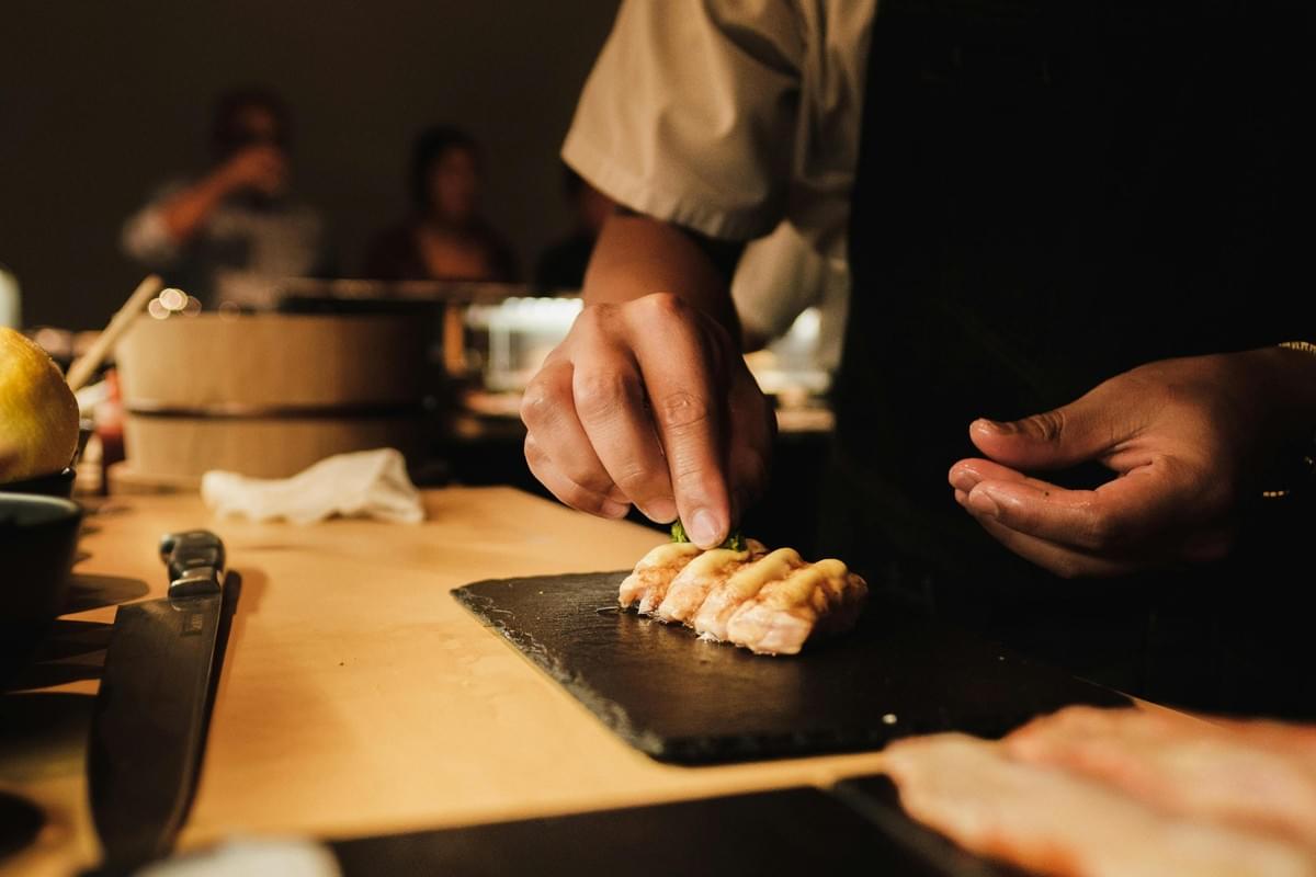 This close-up captures a chef's hands meticulously garnishing a row of sauce-topped sushi or sashimi pieces arranged on a dark serving slate. Warm lighting highlights the wooden countertop and sharp knife in the foreground, while the blurred background suggests a busy, intimate dining atmosphere.