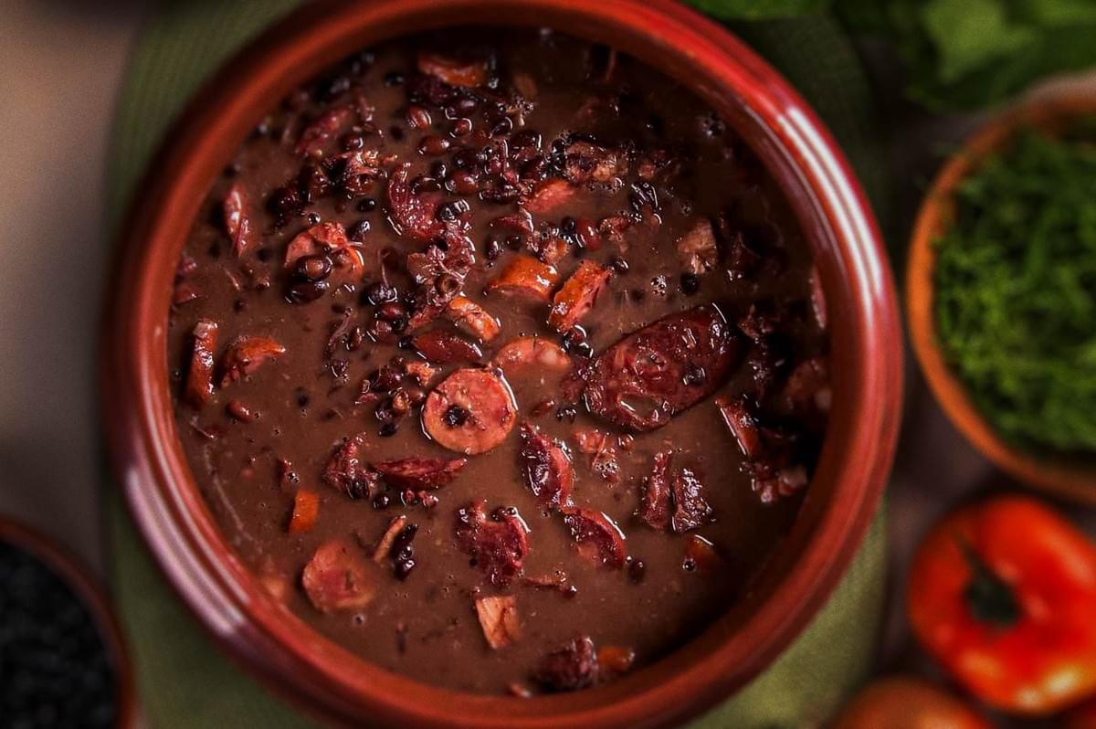 Brazilian feijoada stew in rustic clay pot with black beans, sausage slices, and assorted pork and beef cuts, served with fresh greens and tomatoes.