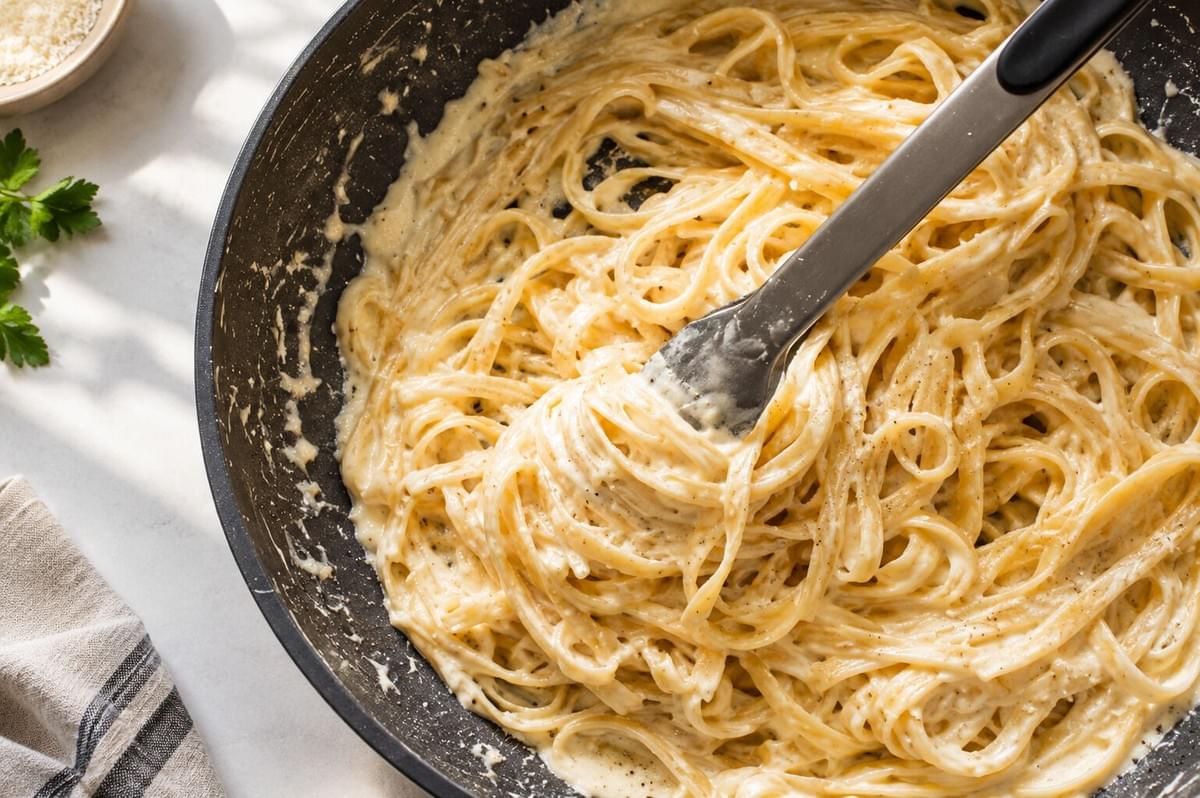 Tongs tossing fettuccine pasta in a creamy white sauce inside a skillet.