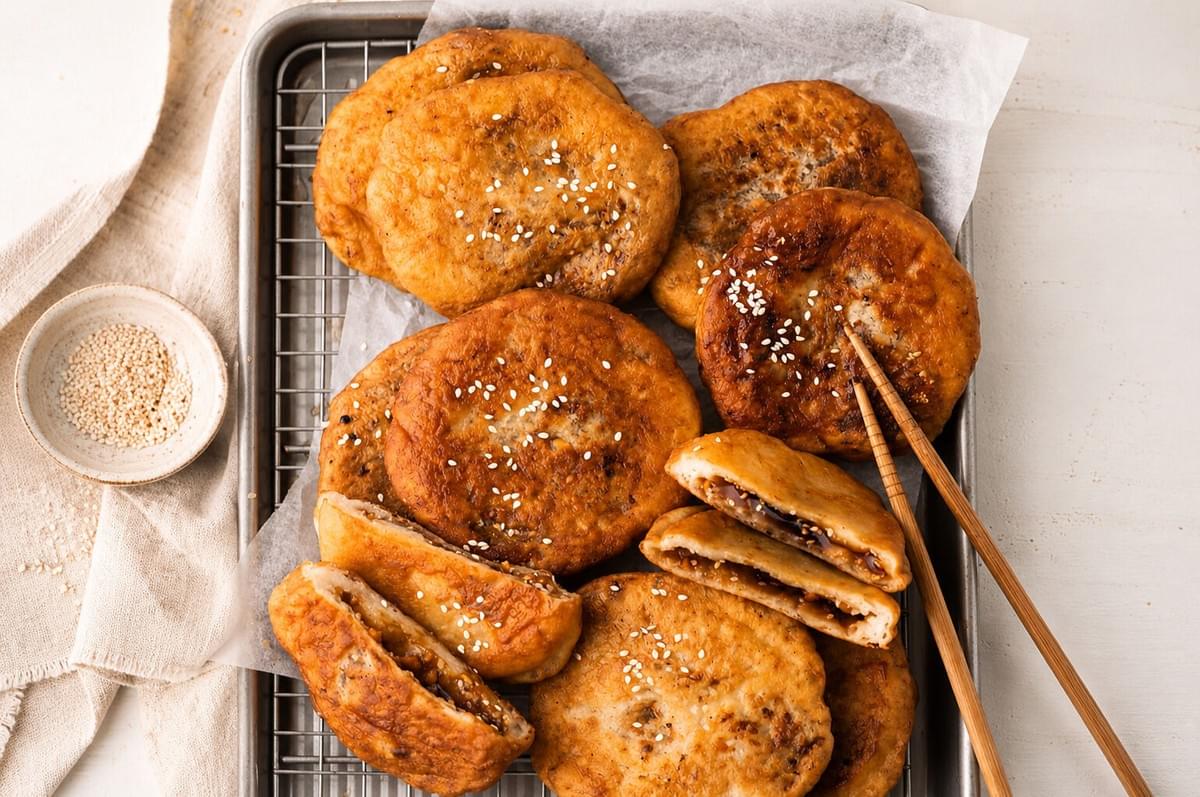 Golden-brown hotteok pancakes sprinkled with sesame seeds cooling on a wire rack with chopsticks.