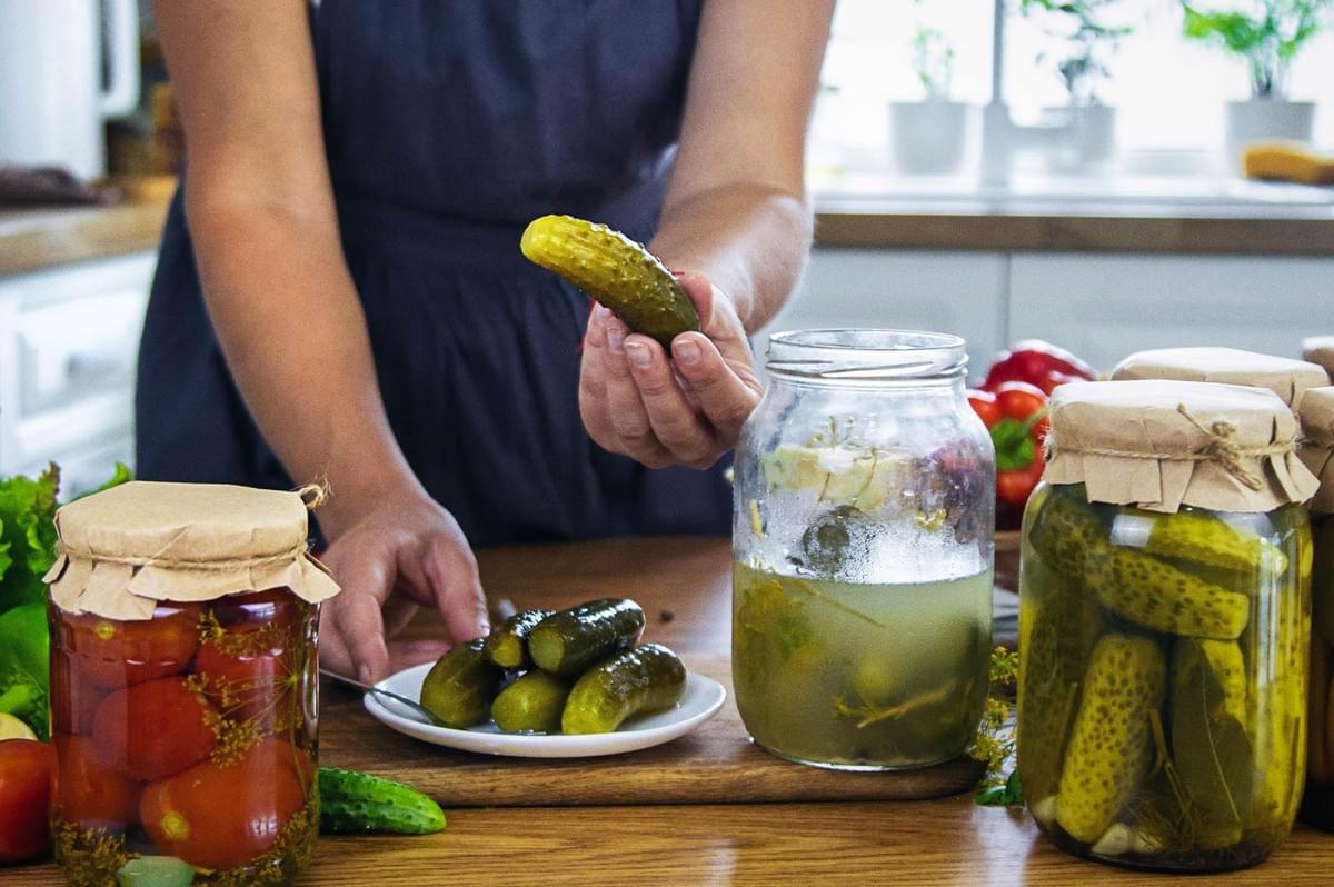 A person standing at a kitchen counter holding a pickle above a plate, surrounded by jars of homemade pickled vegetables including cucumbers and tomatoes.