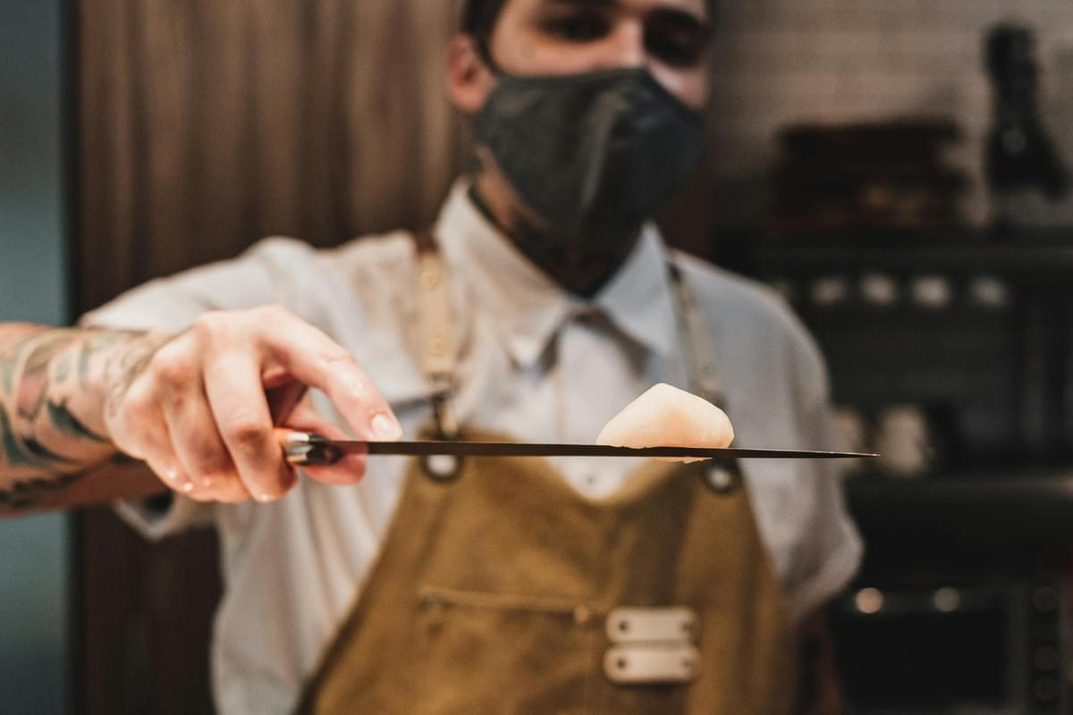 This image showcases a masked chef with tattooed arms presenting a piece of food balanced on the flat side of a knife blade. The shallow focus draws attention to the stability of the chef's hand and the texture of the ingredient, blurring the kitchen background to highlight professional precision.