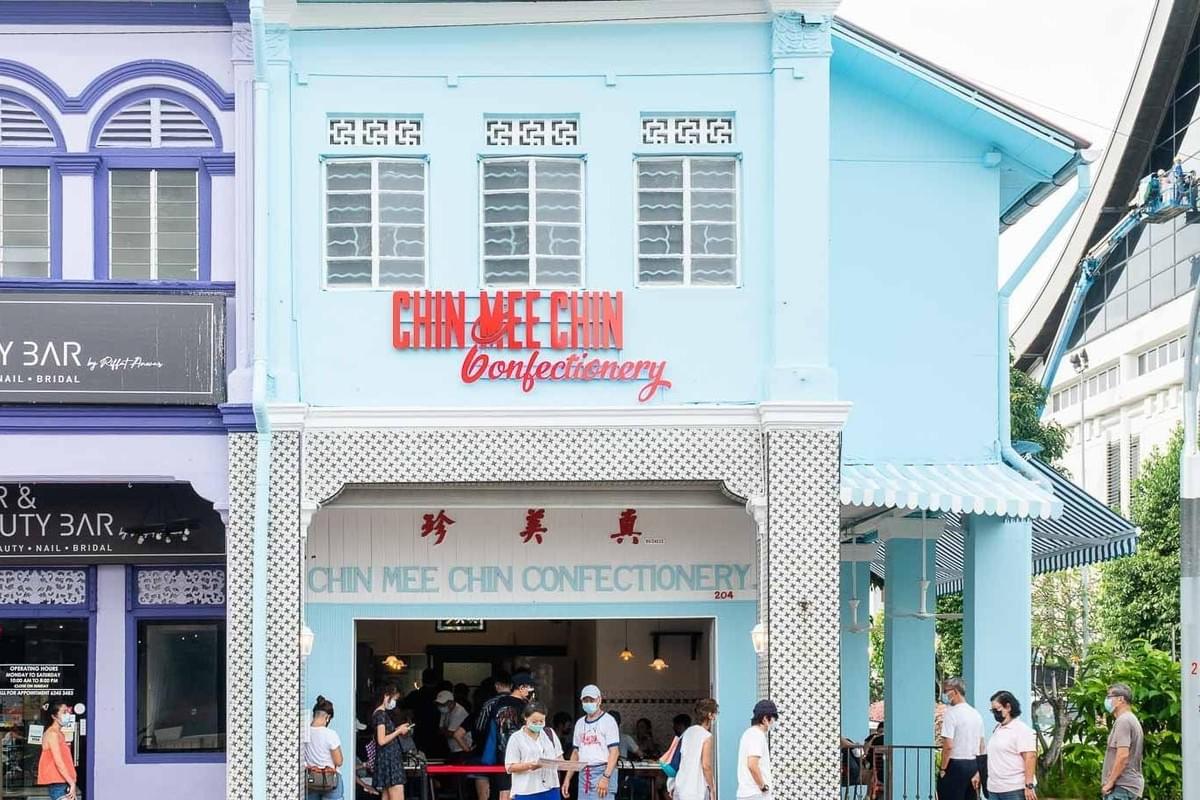 The image features the light-blue facade of Chin Mee Chin Confectionery, a traditional shophouse with red signage and decorative patterned tiling. Customers are seen gathering near the open entrance and outdoor seating area, which is partially shaded by a blue-and-white striped awning.
