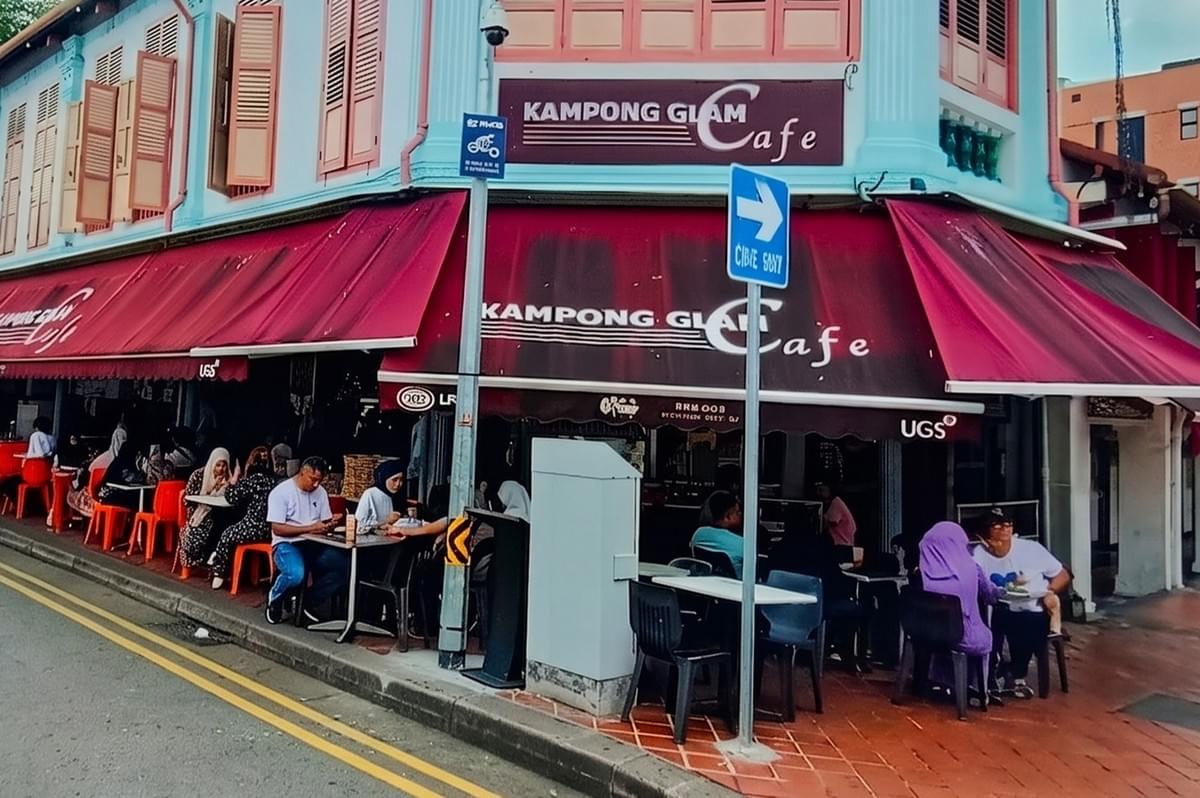 Exterior of Kampong Glam Cafe, a corner restaurant with outdoor seating and red awnings on a city street.