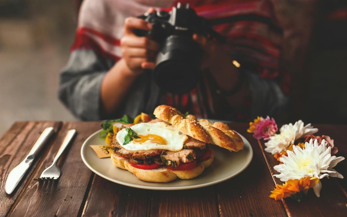 A person holds a DSLR camera positioned to take a photo of an open-faced breakfast sandwich on a wooden table. The sandwich is served on a braided bun topped with sliced meat, tomatoes, and a sunny-side-up egg, flanked by cutlery and decorative flowers.
