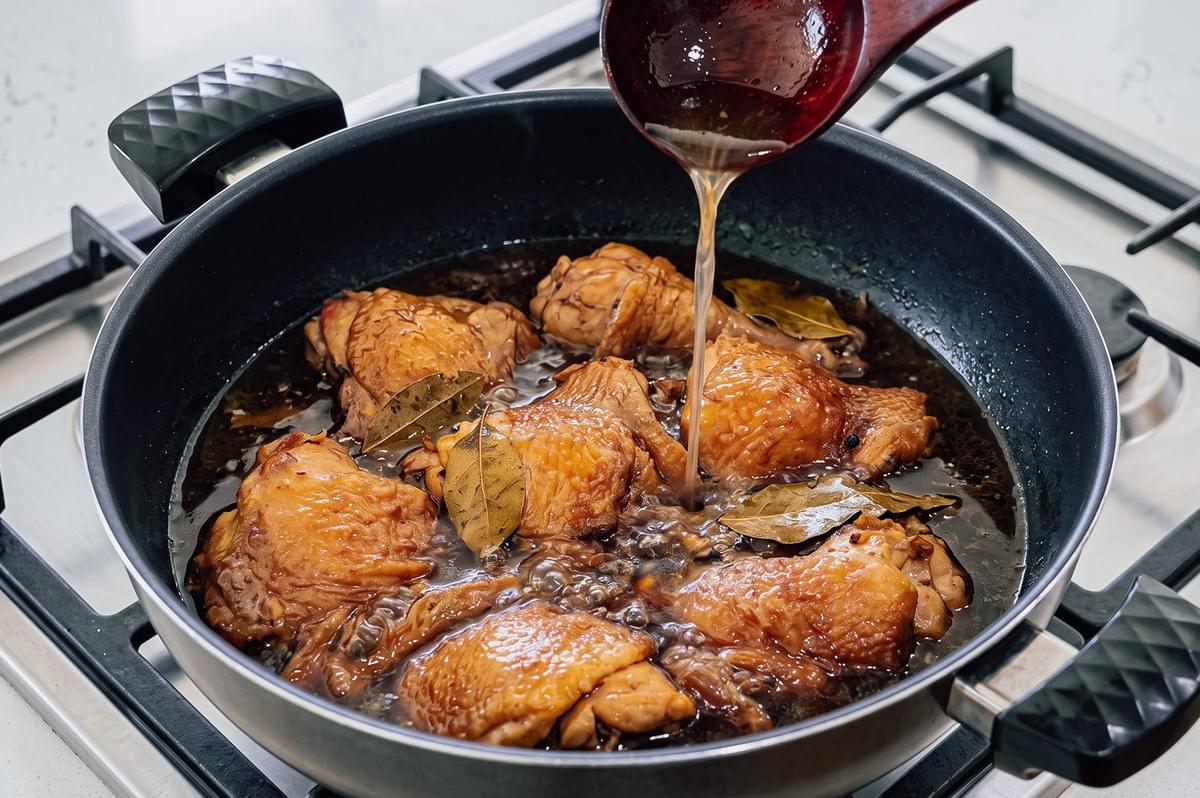 High-angle view of Filipino chicken adobo simmering in a pan as vinegar is poured into a savory soy sauce broth with garlic, bay leaves, and peppercorns during the traditional cooking process.