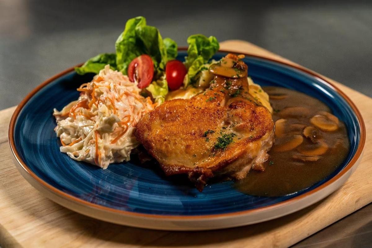 A golden-brown, pan-seared chicken chop sits on a blue plate, served with a side of coleslaw, fresh greens, and cherry tomatoes. A rich, savory mushroom gravy is generously poured next to the chicken and over a bed of mashed potatoes.