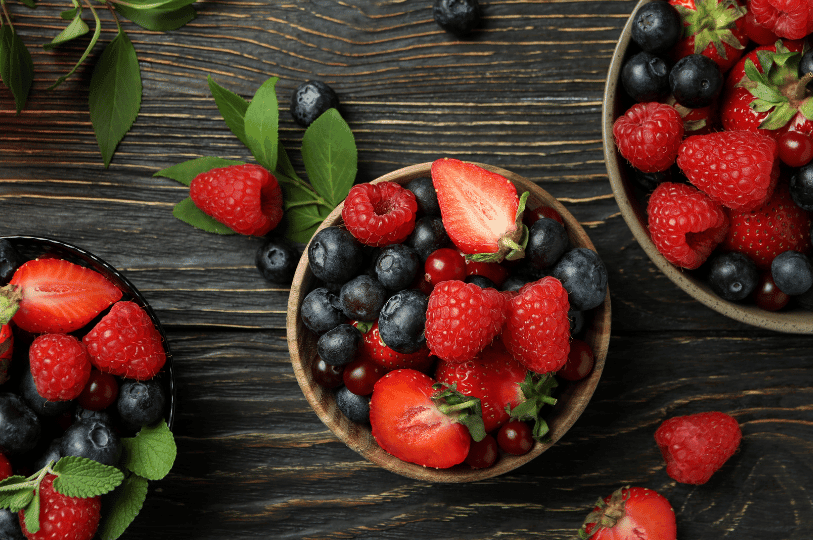 An overhead view of three small wooden bowls filled with sorted strawberries, blueberries, and raspberries on a dark wood background, demonstrating the preparation and sorting phase of preservation.