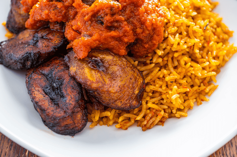 A beautifully styled ceramic bowl filled with savory Jollof rice, tender chunks of beef stew, and a side of sweet, ripe plantains resting on a textured wooden table.