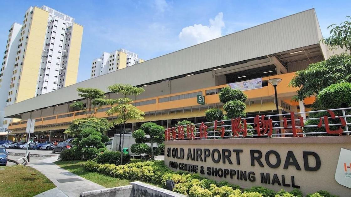 This image shows the exterior of the 51 Old Airport Road Food Centre & Shopping Mall in Singapore, a prominent two-story building featuring yellow and orange accents. The foreground displays the center's sign and manicured greenery.