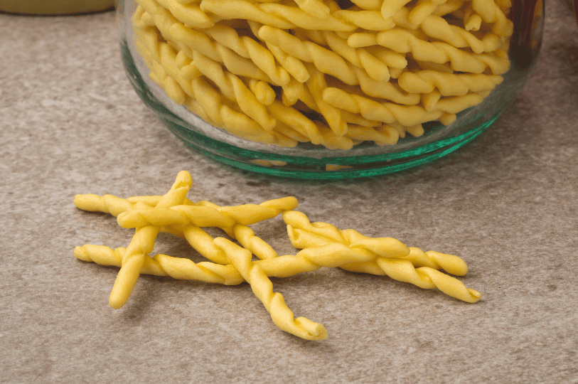 Pieces of uncooked trofie, a short, tightly twisted pasta typical of Liguria, resting on a counter near a glass jar filled with more of the same shape.