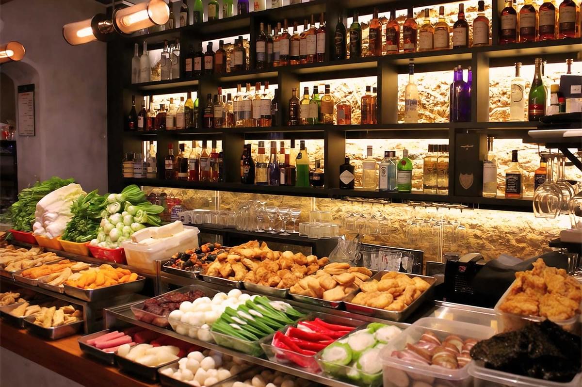 A vibrant bar scene with shelves of various liquor bottles against a warmly lit wall. In the foreground, colorful vegetables and fried snacks are neatly arranged on a counter.