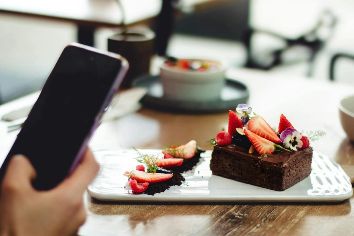 A person holds a smartphone over a wooden table to take a photo of a plated chocolate dessert. The treat is elegantly garnished with fresh strawberries, raspberries, and edible flowers on a white rectangular plate.