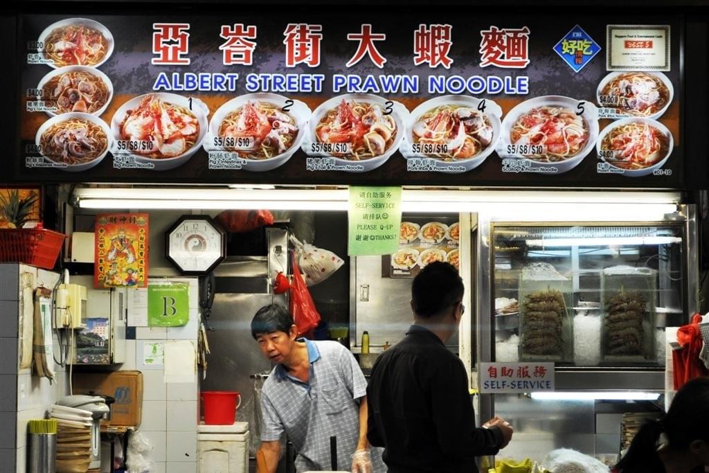 The image shows a hawker stall named Albert Street Prawn Noodle, featuring a large menu board with photos and prices for various prawn noodle dishes. A stall assistant in a blue checkered shirt is visible behind the counter, which displays a "Self-Service" sign and a refrigerator containing ingredients.