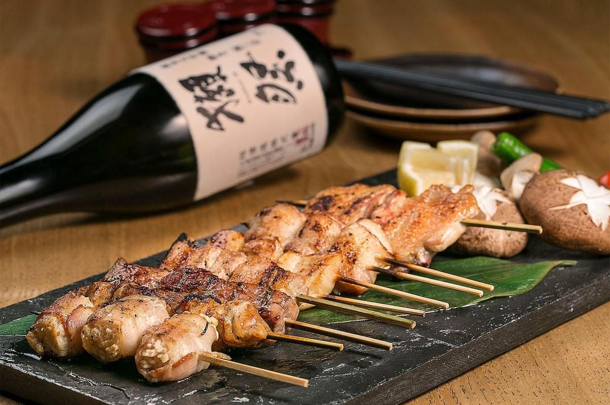 A line of grilled yakitori skewers sits arranged on a slate platter alongside shiitake mushrooms and lemon wedges. In the background, a dark glass sake bottle lies on a warm wooden table next to chopsticks and small dishes.