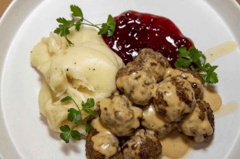 A detailed close-up of Swedish meatballs heavily coated in a smooth, light-brown cream sauce, accompanied by buttery mashed potatoes, lingonberry preserves, and fresh parsley sprigs.
