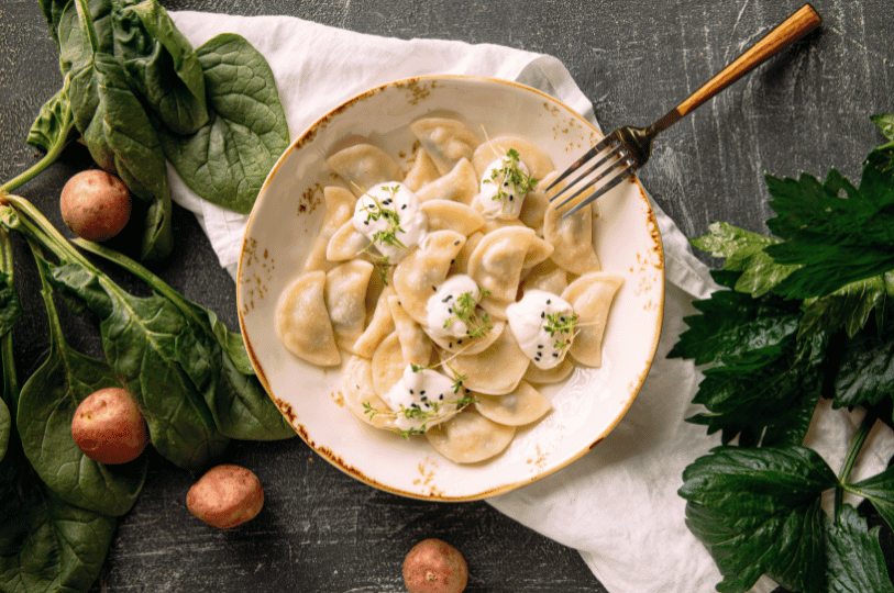 A flat-lay view of a plate of traditional pierogi topped with dollops of sour cream, black sesame seeds, and microgreens, surrounded by raw spinach and potatoes.