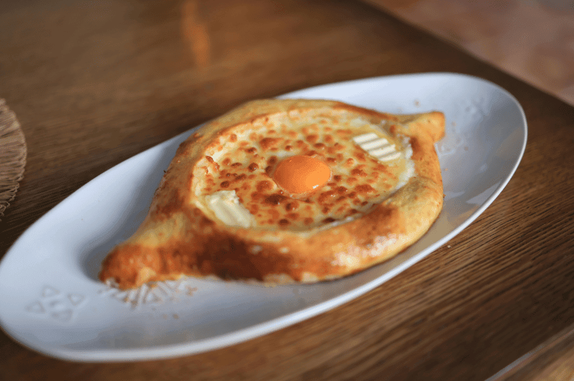 A wide-angle perspective of an Adjaruli Khachapuri resting on an elegant white plate. The lighting highlights the glistening butter and the vibrant orange yolk sitting atop a bed of toasted, browned cheese.