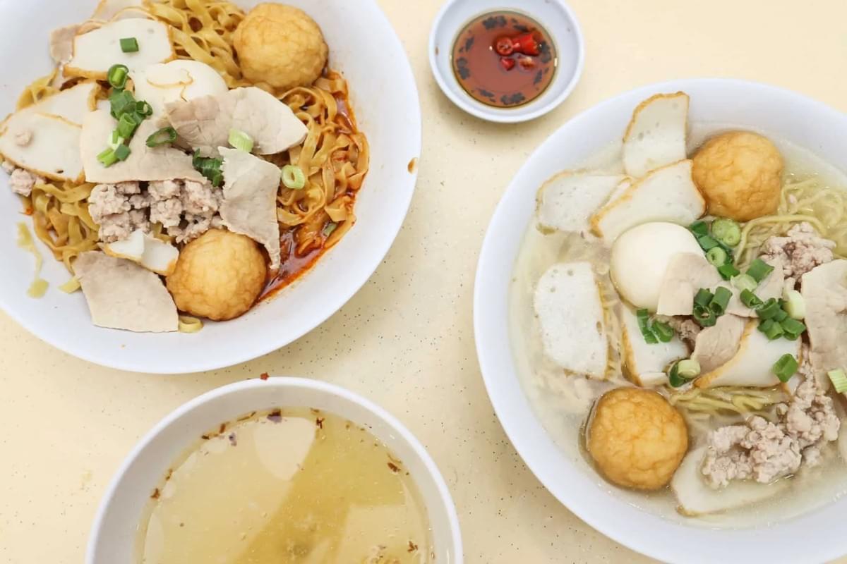 This overhead shot displays two bowls of noodles topped with fishballs, sliced fish cakes, and minced meat, accompanied by a small side of chili sauce. One bowl features dry noodles tossed in a savory sauce, while the other serves the ingredients in a light, clear broth.