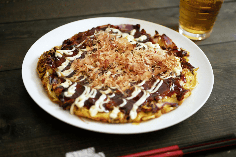 A freshly cooked okonomiyaki topped with creamy mayonnaise, rich brown sauce, and fluttering bonito flakes, served on a white plate with chopsticks—capturing the irresistible allure of Japan’s iconic savory pancake.