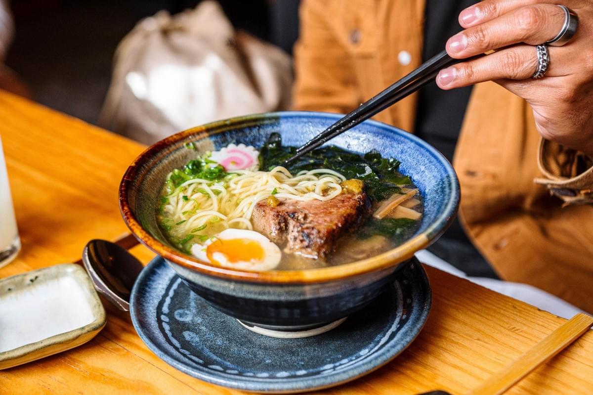 In the image, a vibrant bowl of ramen sits on a wooden table, filled with noodles, broth, a soft-boiled egg, sliced pork, seaweed, and a narutomaki fish cake. A person wearing a tan jacket uses black chopsticks to lift some of the contents from the blue and brown ceramic bowl.