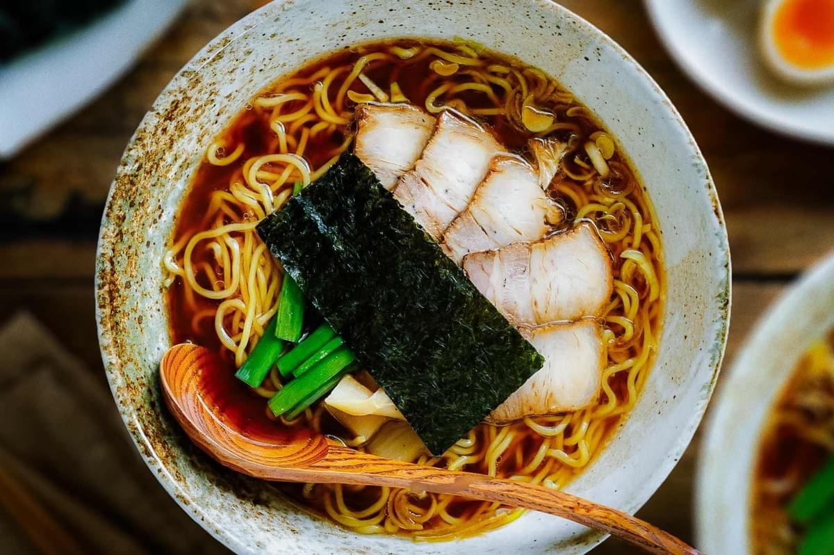 Bowl of ramen with noodles, pork slices, seaweed, bamboo shoots, and green onions in broth, wooden spoon resting inside.
