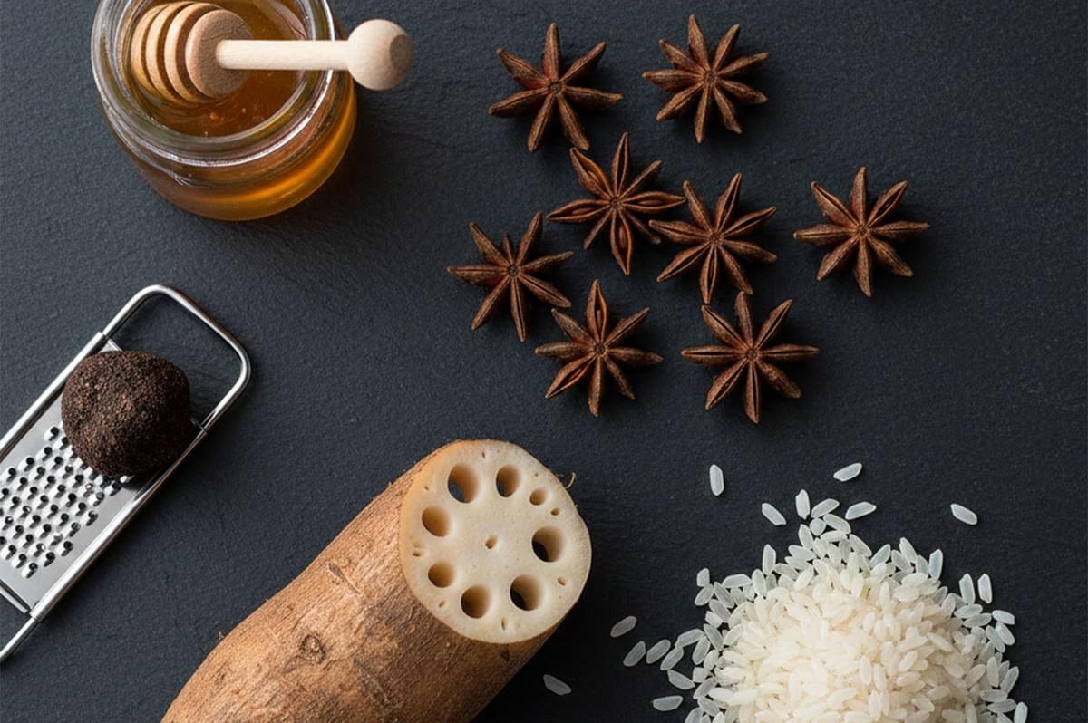 Top-down view of gourmet risotto ingredients on a dark slate background, featuring raw lotus root, a black truffle with a grater, star anise, a jar of honey, and uncooked arborio rice.