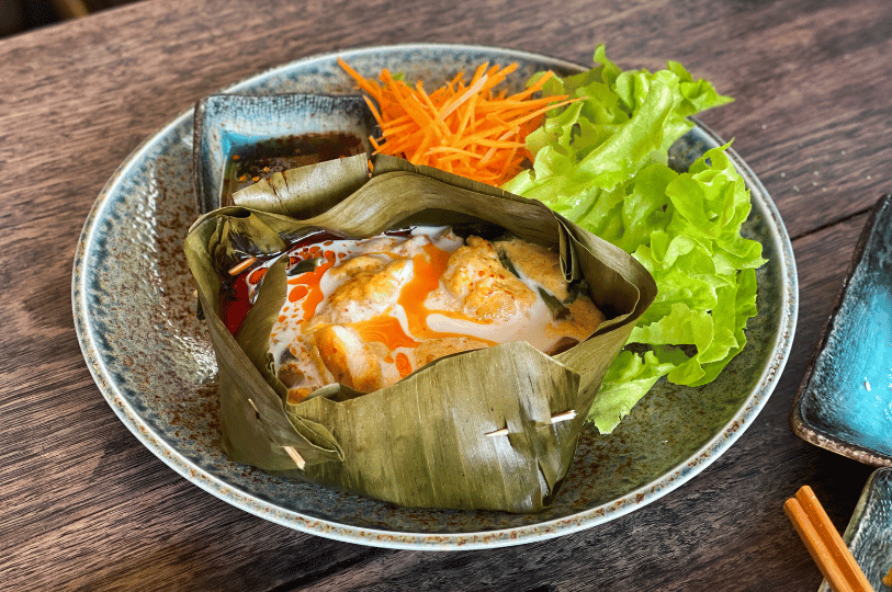 A beautifully plated Cambodian fish amok in a round banana leaf cup, showcasing the golden curry topped with a dollop of thick coconut cream, a fresh cilantro leaf, and slivers of red pepper.