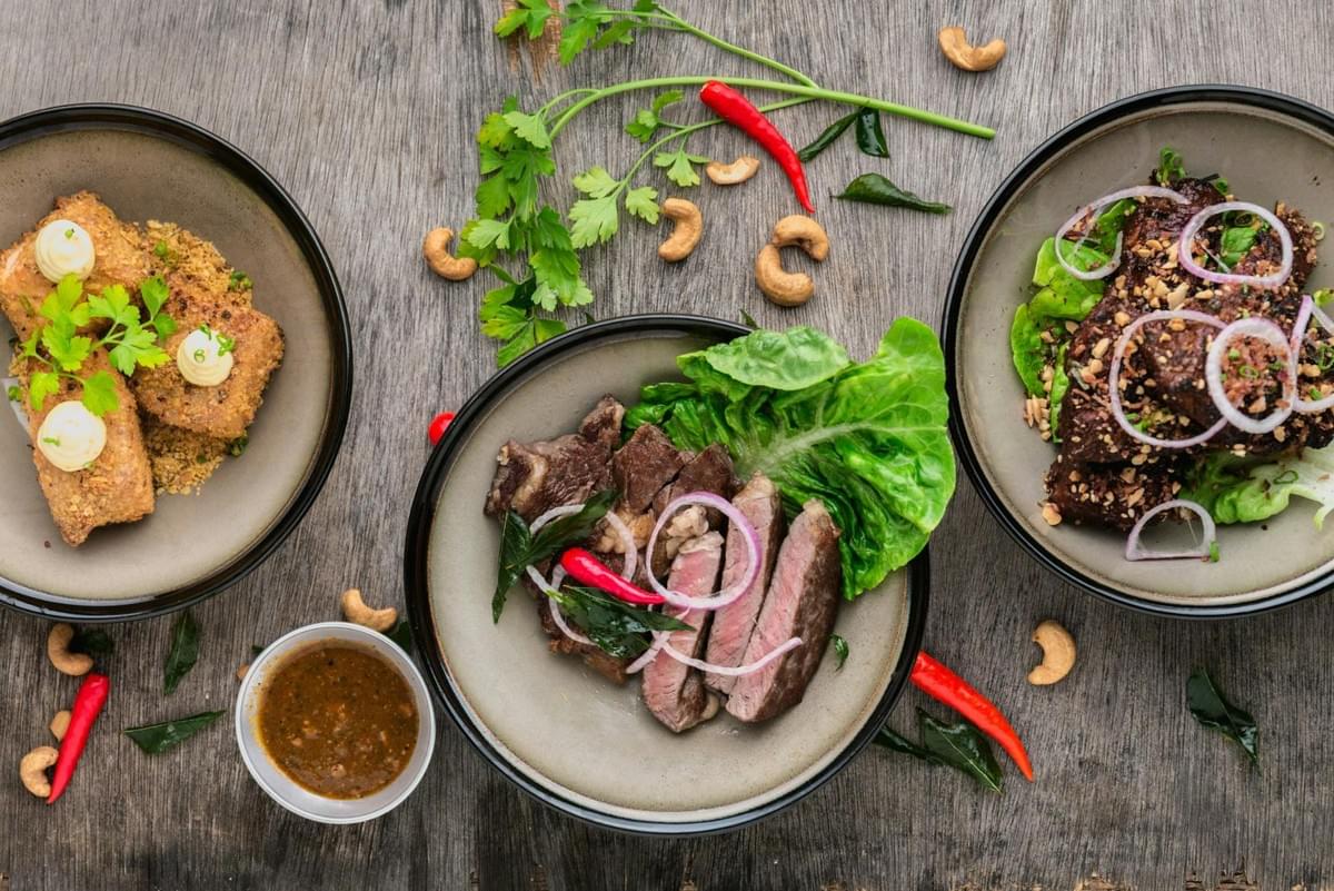 An overhead view features three dark bowls filled with various savory dishes arranged across a rustic wooden table alongside a small bowl of dipping sauce. The meal includes sliced steak garnished with onion rings and red chilies, crispy fried rolls with creamy dollops, and dark grilled meat topped with herbs and crushed nuts.