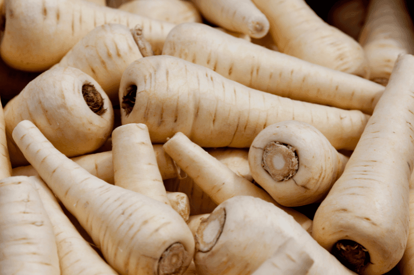 A rustic overhead shot of raw, tapered parsnips with some dirt still on their skins, resting on a dark wooden surface to highlight their natural, earthy origin.