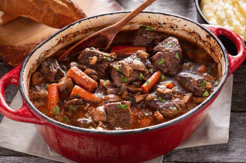 A rustic red enameled cast-iron pot filled with Beef Bourguignon, a traditional Burgundian stew. The rich, dark wine sauce coats tender chunks of beef, bright orange carrot rounds, and whole button mushrooms.