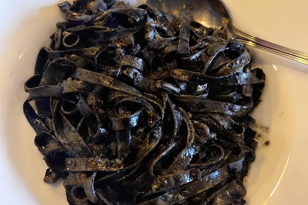 Close-up of a serving of black squid ink pasta on a white plate with a metal spoon.
