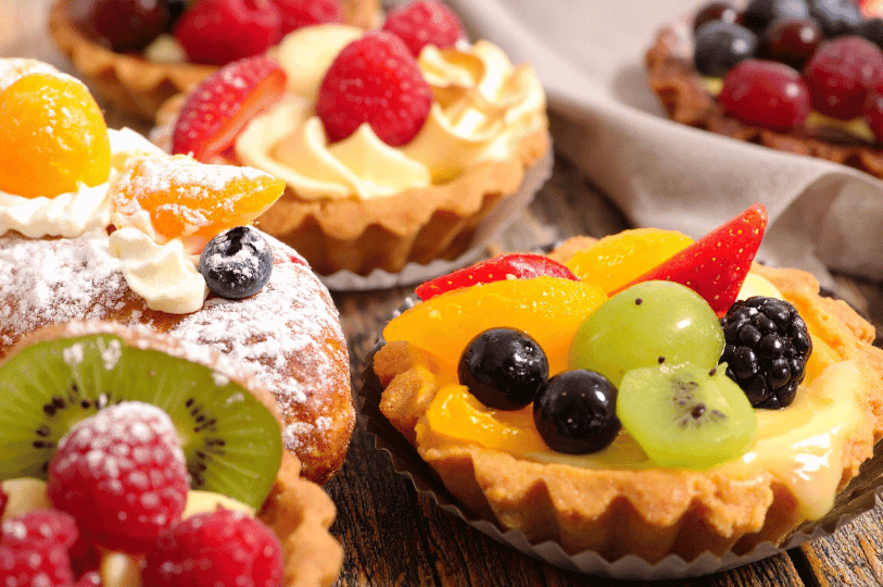An assortment of fruit tarts and pastries topped with berries, kiwi, and peach slices rests on a rustic wooden surface, their powdered sugar and whipped cream accents celebrating the diversity of tart styles and seasonal toppings.