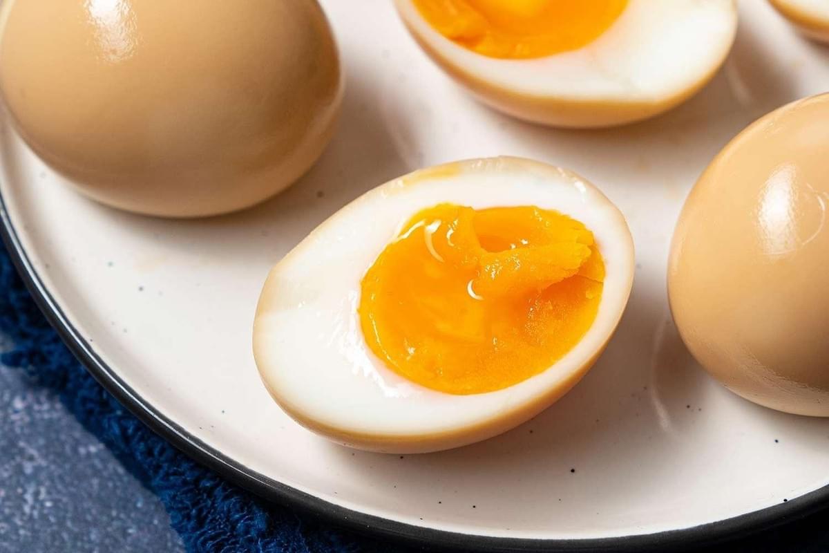 A plate of soy-marinated ramen eggs, with one sliced in half to reveal a jammy, bright yellow yolk. Whole light-brown marinated eggs surround the half piece on a ceramic plate.