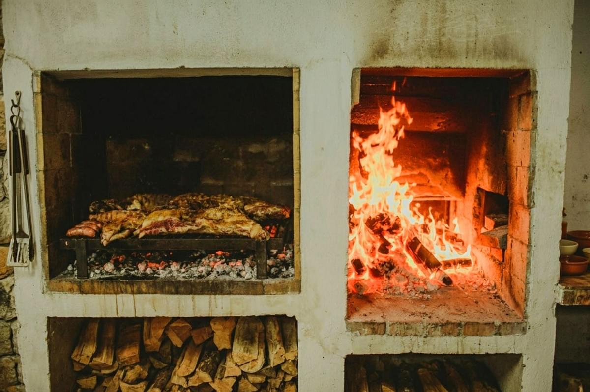 Traditional Argentine parrilla with meat grilling over embers in left chamber and active wood fire in right chamber, firewood stored below