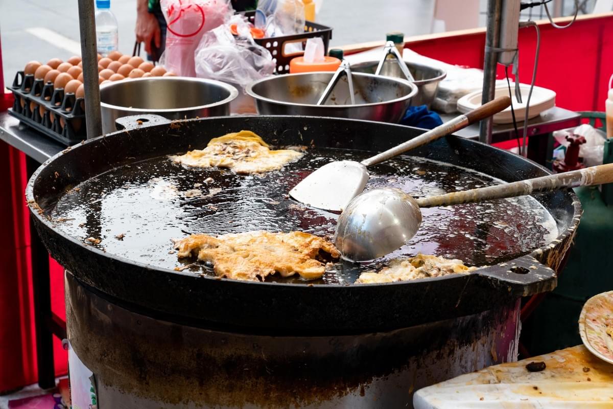 Large, crispy omelets fry in a shallow, oversized metal wok filled with bubbling oil, with sturdy metal spatulas resting inside the pan. The surrounding street food stall is stocked with a carton of fresh brown eggs and stainless steel bowls containing ingredients ready for preparation.