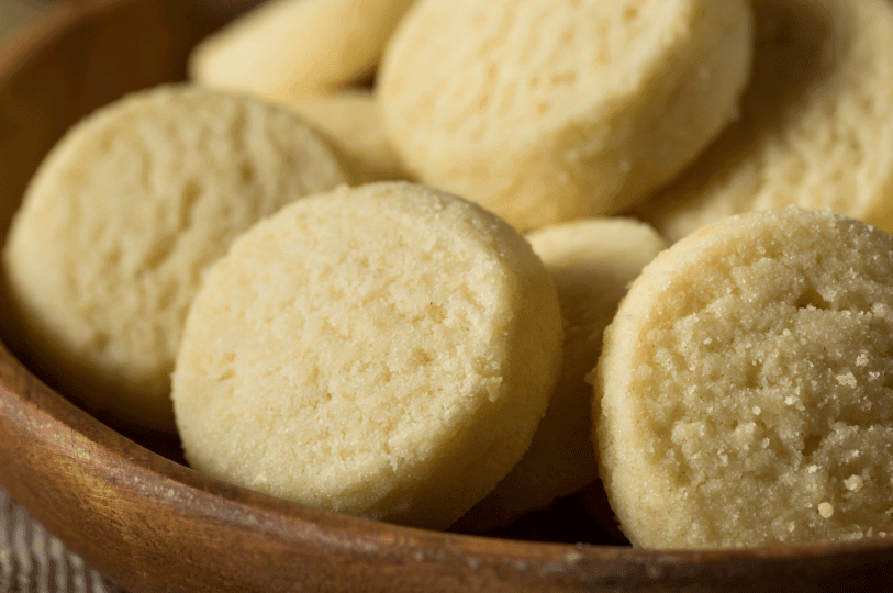 A close-up, angled shot of thick, round shortbread cookies in a wooden bowl, highlighting their pale golden color and slightly porous, crumbly texture.