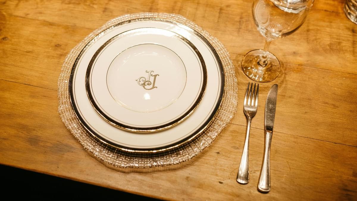 This overhead shot features an elegant place setting on a rustic wooden table, consisting of a dinner plate and a smaller salad plate both with gold and white trim. To the right of the plates, a silver fork and knife are neatly placed next to a stemmed wine glass.