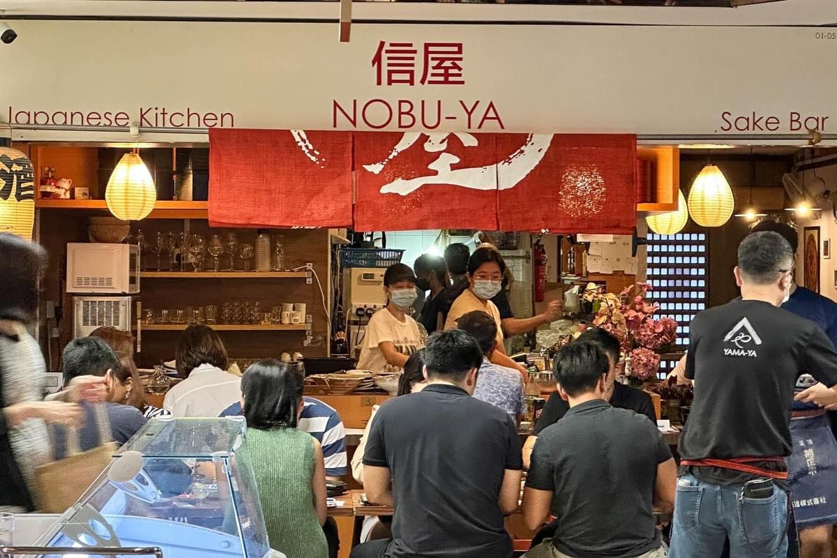 Patrons sit at a long counter facing the open kitchen of Nobu-Ya, a Japanese restaurant and sake bar. Staff members wearing masks work behind the counter under warm, glowing lanterns and red fabric banners.