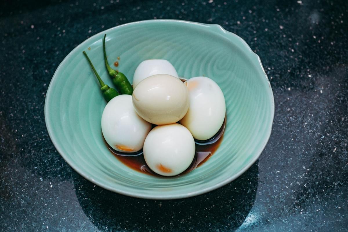 A light green ceramic bowl rests on a dark speckled surface, filled with peeled hard-boiled eggs sitting in a savory brown sauce. Two whole green chili peppers are tucked along the side of the bowl as a garnish.