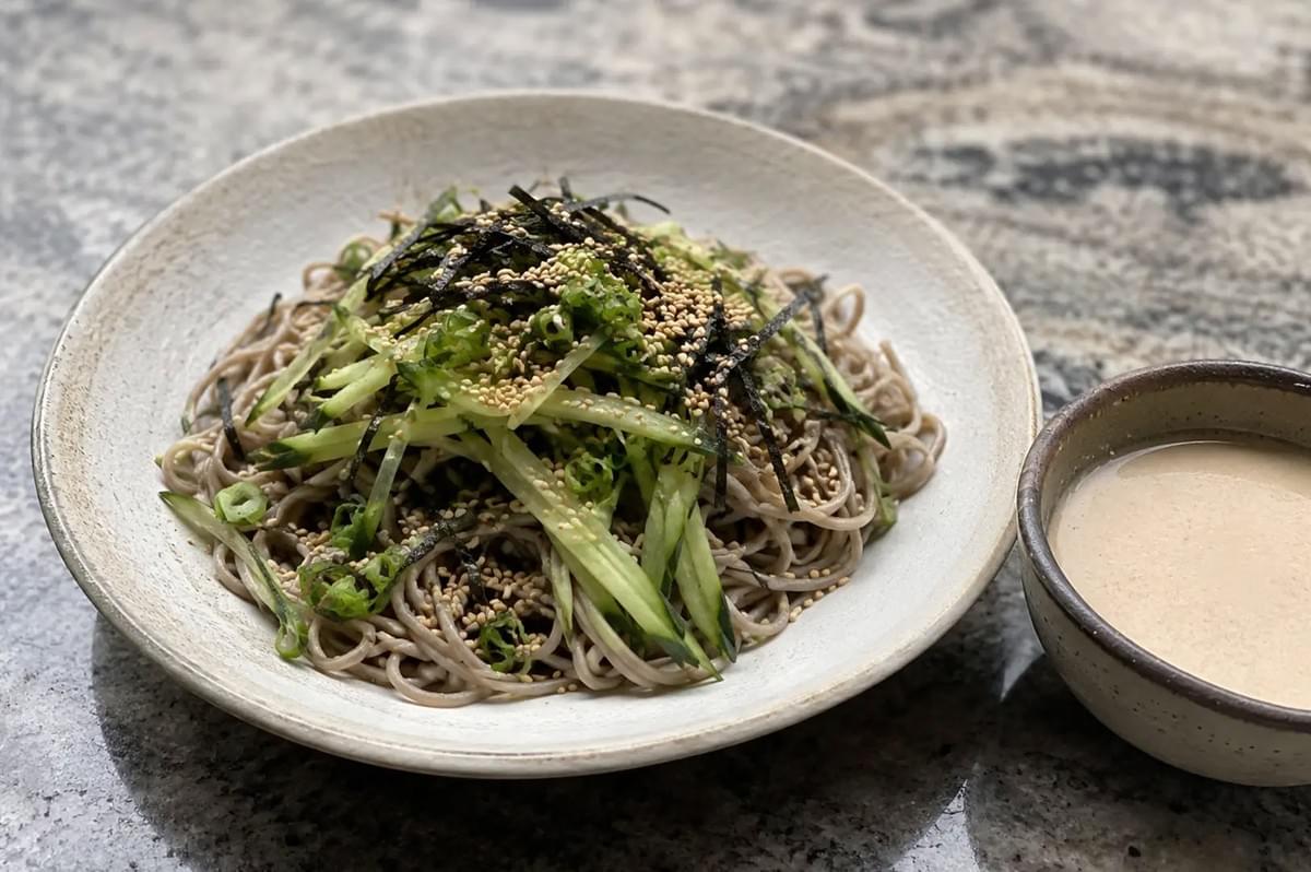 A wide, shallow beige ceramic bowl filled with cold soba noodles sits on a dark gray granite countertop. The buckwheat noodles are mounded in the center, generously topped with a mountain of thinly julienned green cucumbers, finely chopped scallions, toasted white sesame seeds, and shreds of dark green nori seaweed. To the right of the main dish sits a small, matching ceramic bowl containing a smooth, light beige sesame-tahini mentsuyu sauce, ready for dipping or pouring.