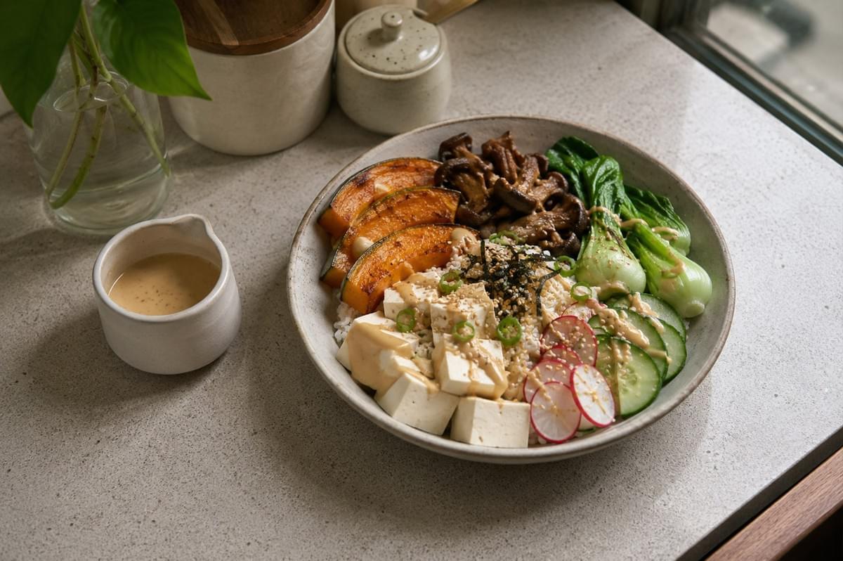 A top-down, slightly angled shot of a beautifully plated vegetarian Japanese-style bowl resting on a light-colored countertop. The bowl features a base of white rice topped with cubed tofu, roasted pumpkin slices, sautéed mushrooms, blanched bok choy, thinly sliced radishes, and fresh cucumber rounds, all drizzled with a light-colored sesame-based dressing and garnished with sesame seeds and nori strips. Beside the main bowl sits a small matching ceramic pitcher filled with the same creamy dressing, and a clear glass vase with greenery is visible in the corner.