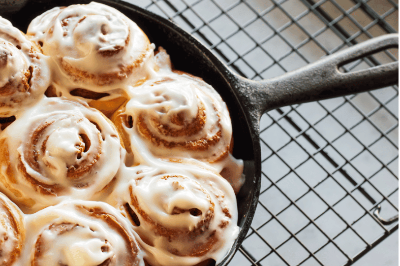 Golden-brown cinnamon rolls drizzled with gooey icing, served warm in a black cast-iron skillet next to a cooling rack.