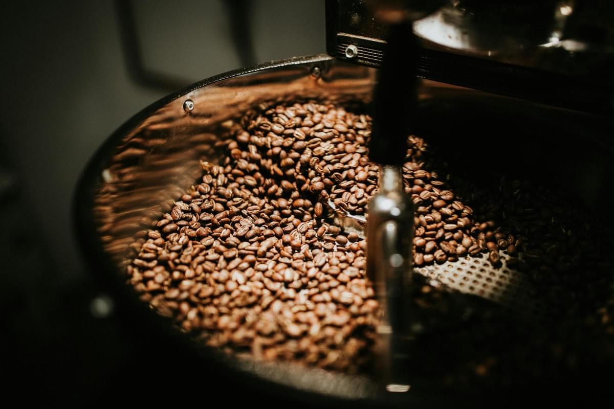 Inside the metallic drum of an industrial roaster, a large quantity of coffee beans is captured during the cooling process. The dramatic, warm lighting highlights the texture and rich brown color of the beans, creating a sharp contrast with the dark, mechanical surroundings.