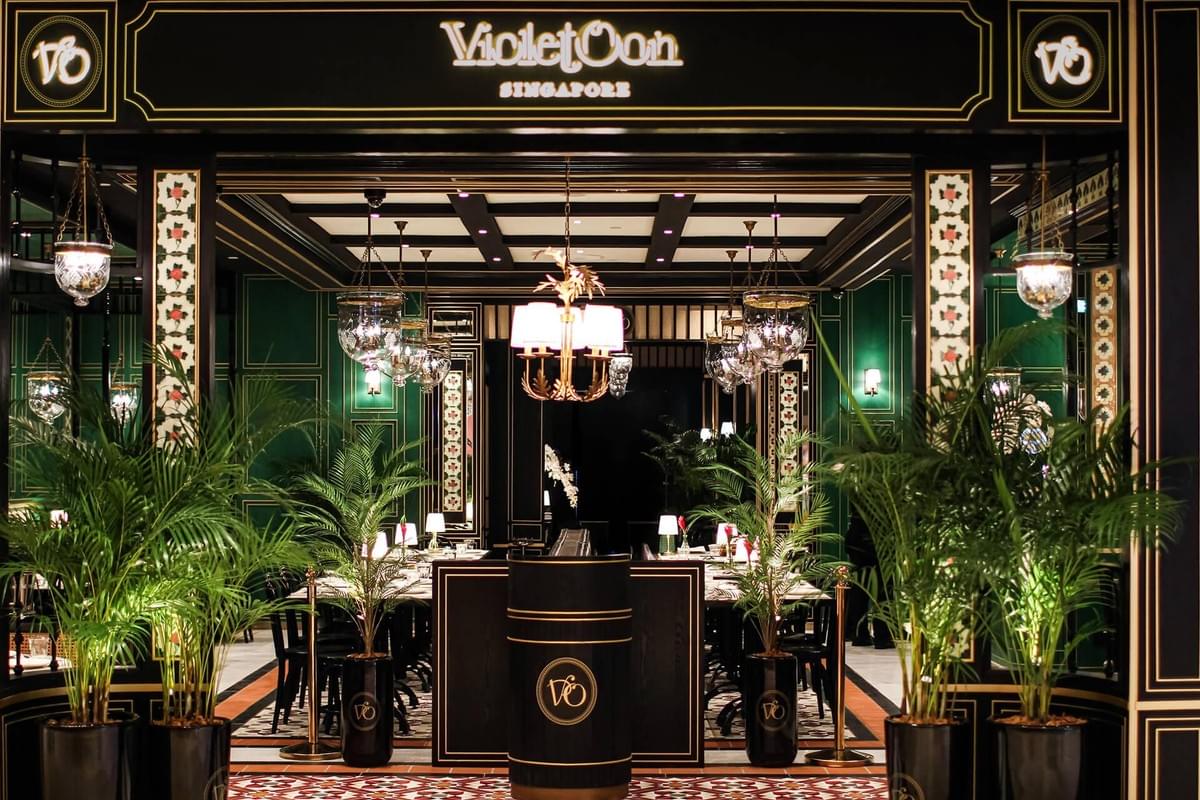  Gemini said This image showcases the elegant entrance of Violet Oon Singapore, a restaurant featuring a sophisticated black and gold storefront with lush green potted palms. The interior is illuminated by ornate glass chandeliers and warm table lamps, highlighting a rich color palette of emerald green walls and intricate floral tile flooring.